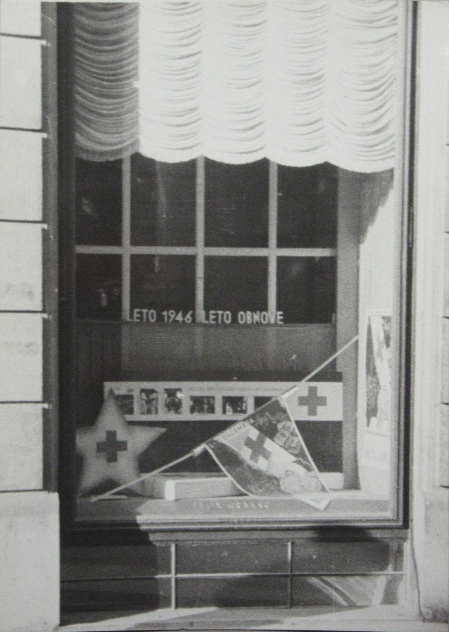 Slika 3: V vseslovenskem »Tednu Rdečega križa« so s prireditvami,
                        zbiranjem denarnih in materialnih sredstev na manifestativen način, kot je
                        okrasitev izložb (na fotografiji izložba trgovine z manufakturo v Ljubljani
                        z napisom LETO 1946 LETO OBNOVE), lajšali pogoje za hitrejšo obnovo. Glavni
                        odbor RKS je izdal navodilo, da se »mesto in dežela okrasita v znamenju
                        Rdečega križa in tako privabljata nove člane«. Danes Teden Rdečega križa
                        Slovenije poteka vsako leto od 8. do 15. maja. 