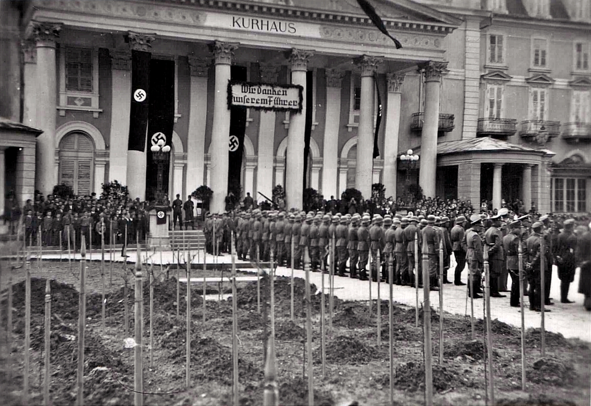 Slika 1: 11. aprila 1942, natanko leto dni po prihodu okupatorja v
                        Rogaško Slatino, je pred Zdraviliškim domom (pod ponemčevalnimi ukrepi
                        preimenovanim v Kurhaus) potekala proslava ob prvi obletnici nemške
                        »osvoboditve« zdraviliškega kraja. Fotografija prikazuje postrojeno četo 60–70
                        nemških vojakov, v ozadju na skrajni levi in desni strani stojijo dečki
                        Hitlerjeve mladine, na sredini ob stebrih so tudi vermani.