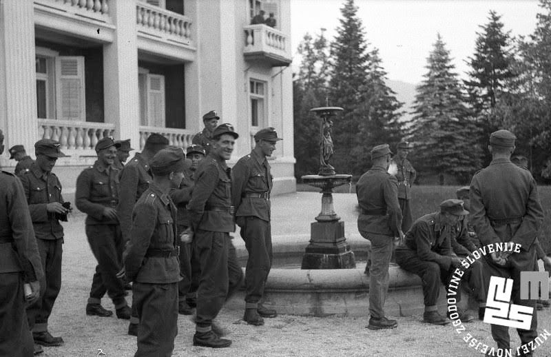 Slika 5: Skupina 19 vermanskih kandidatov, oblečenih v standardno opravo vermanšafta in gorske hlače, na zunanjem dvorišču pred grupno šolo SA v Rogaški Slatini. V ozadju za smrekovim gozdom je razvidno tudi Bočko pogorje, kjer so zlasti v kasnejši fazi vojne operirali lokalni partizanski borci in aktivisti OF.