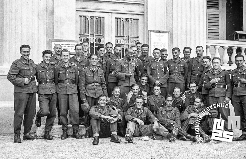 Slika 9: Vermansko moštvo v standardni uniformirani opravi na
                        vojaškopolitičnem usposabljanju v Rogaški Slatini, kjer v sredini tretje vrste
                        stojita tudi predstavnika vodstva šole. Neznana oficirja SA imata na svoji levi
                        roki narokavni trak s svastiko, desni častnik z najvišjim činom na fotografiji
                        pa ima pripeto tudi športno značko SA () oz. športni (vojaški) znak SA s svastiko, vencem in z mečem v ospredju, kar je izkazovalo posameznikove izjemne dosežke. 
